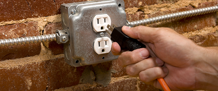 A hand plugging a power cord into an electrical outlet mounted on a brick wall